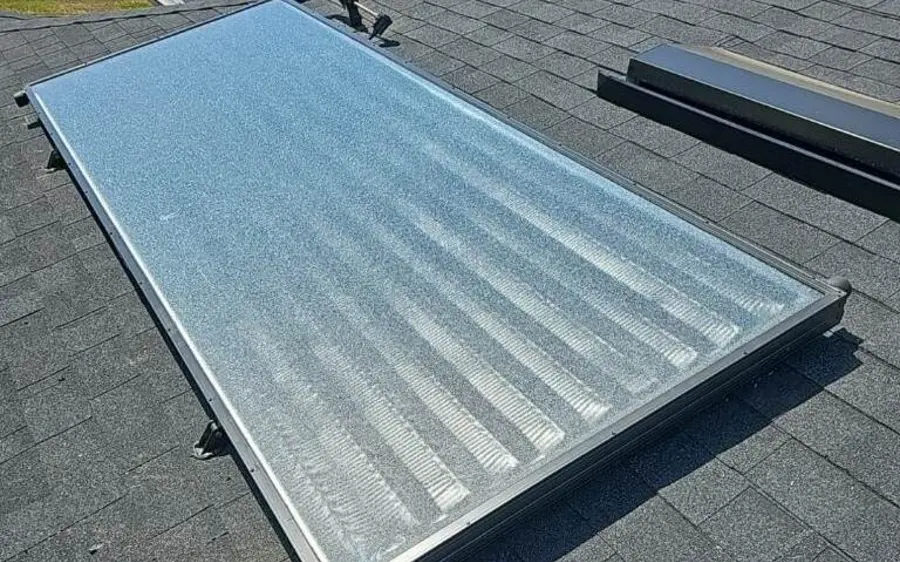 A glazed solar water heating collector mounted flat on a shingle roof, with the supply and return pipes running out of the lower corner.
